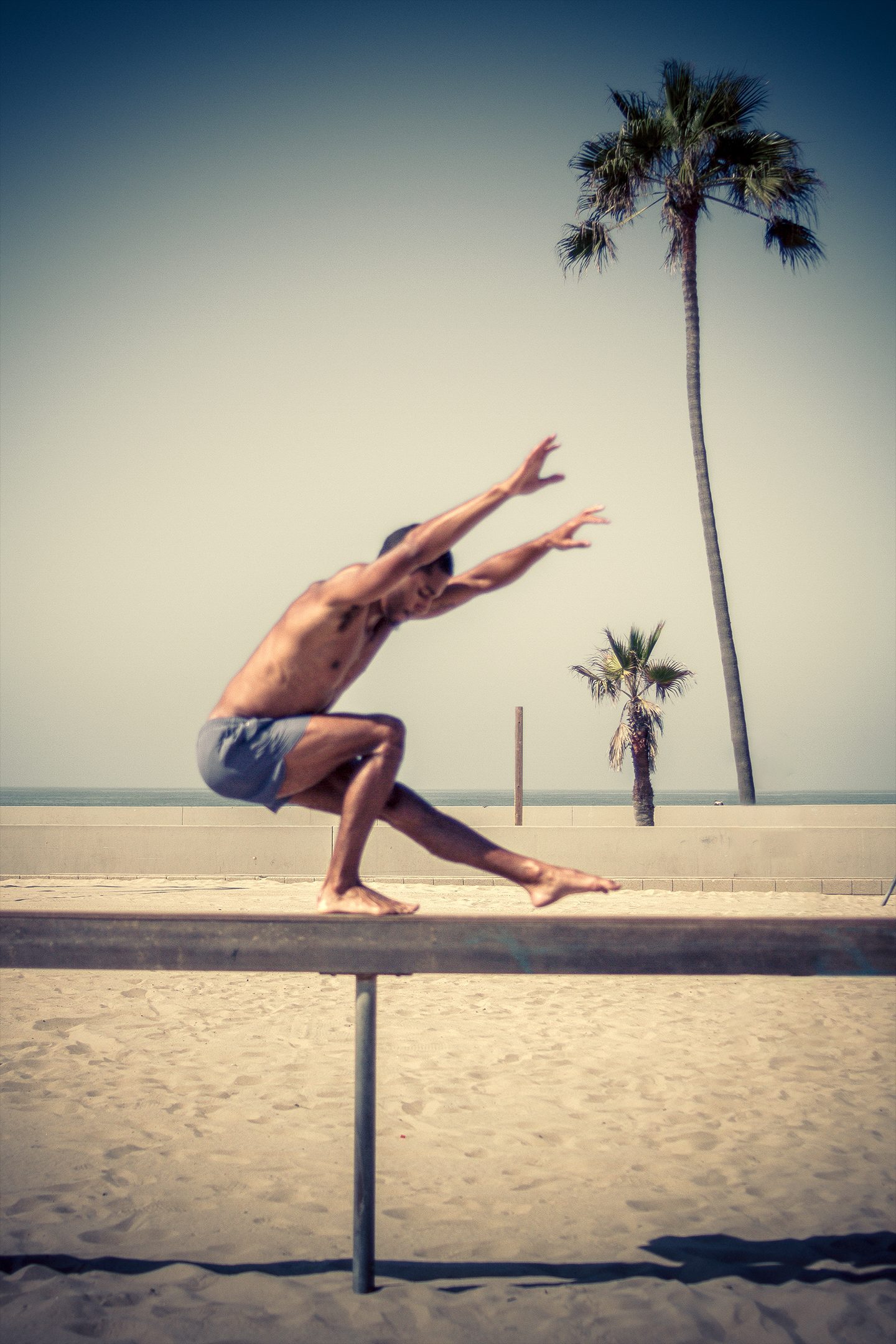 A man balances on a beam on the sand, palm trees behind