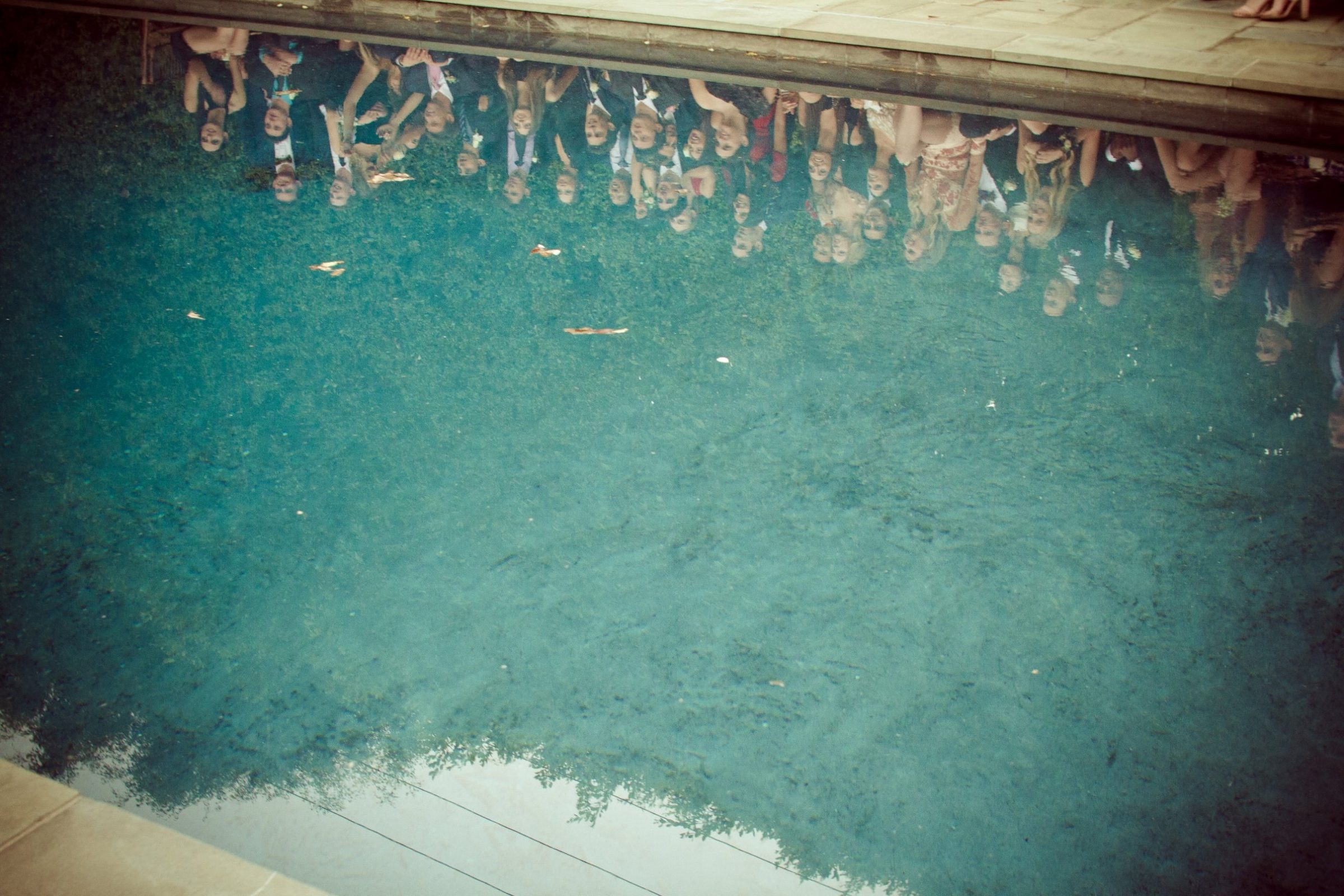 A swimming pool seen from above: a crowd reflected upside down along the far edge, deep teal water, the sky and trees reflected near the corner