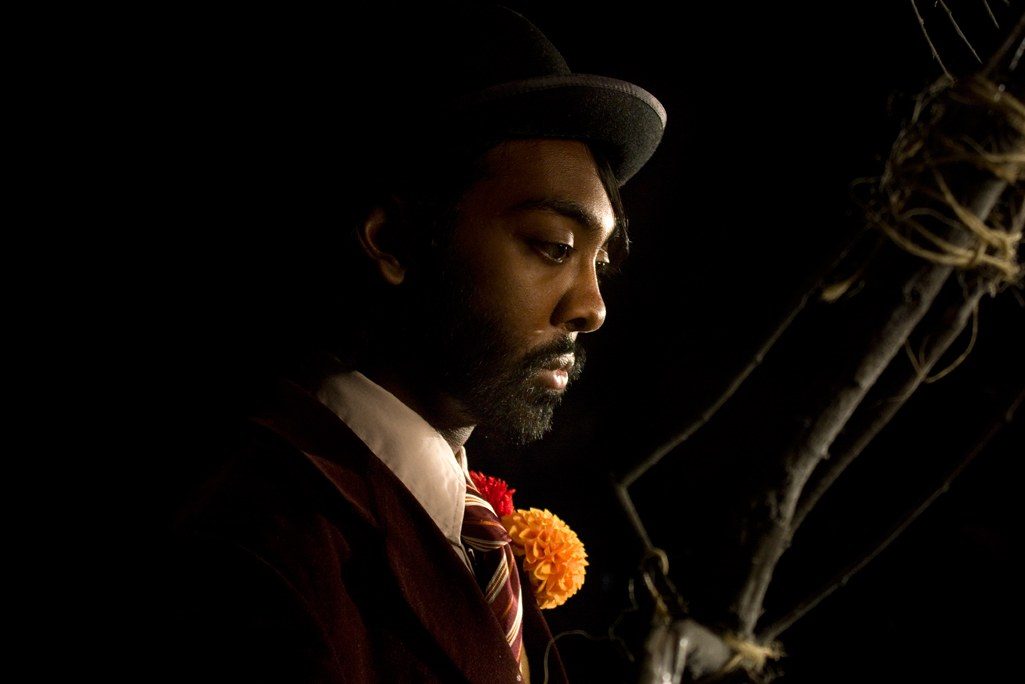 A bearded man in a bowler hat and velvet jacket, lit from one side against black, orange and red flowers at his lapel