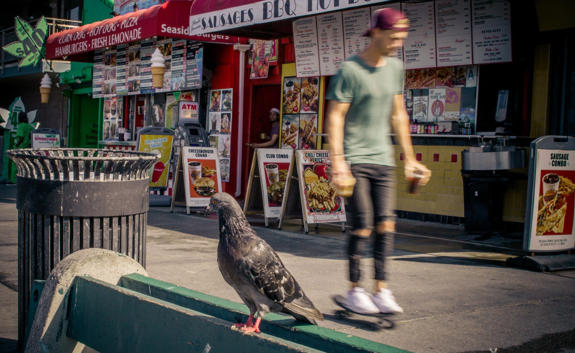 A pigeon on a green bench in sharp focus, a skateboarder blurring past the food stands behind it