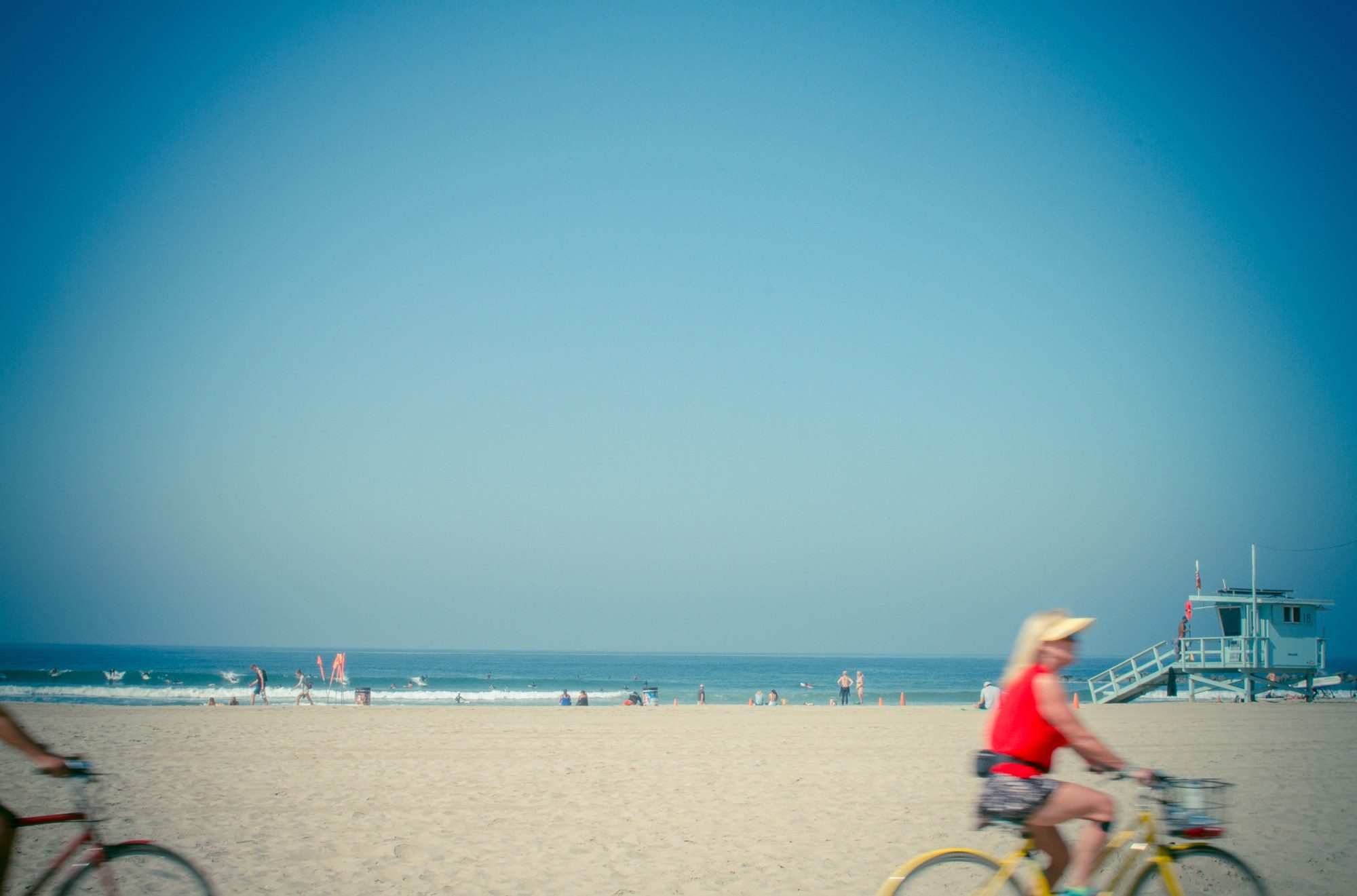 A wide pale blue sky over Venice Beach, two cyclists blurring past in the foreground, a lifeguard tower at right